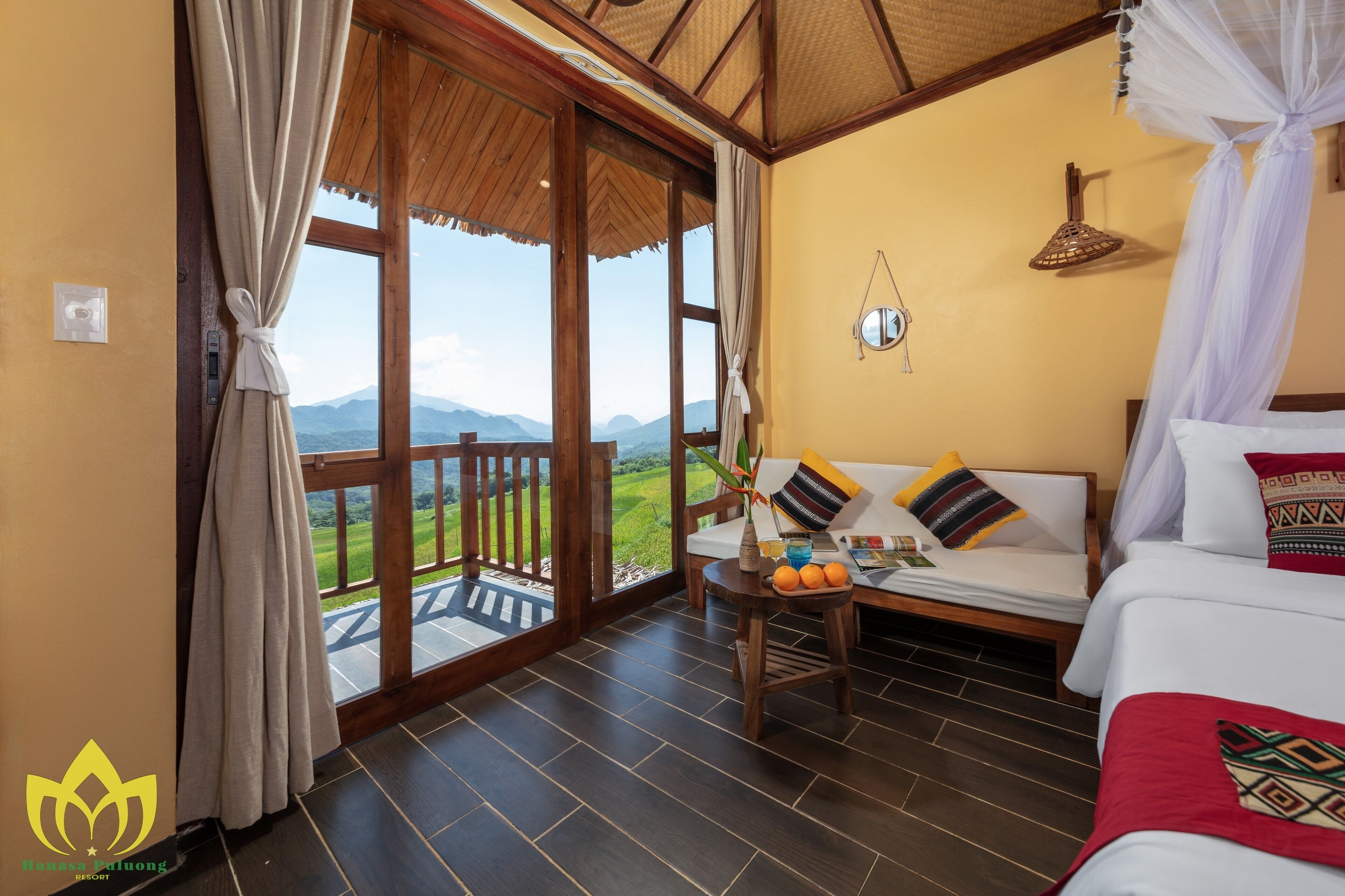 A room with a bed, a sofa, a table with oranges, and a view of mountains through large windows.