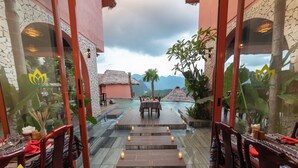 Restaurante con vistas a montañas, que cuenta con un área de comedor envuelta en cristal, mesas y sillas de madera, y una escalera central que conduce a una terraza.