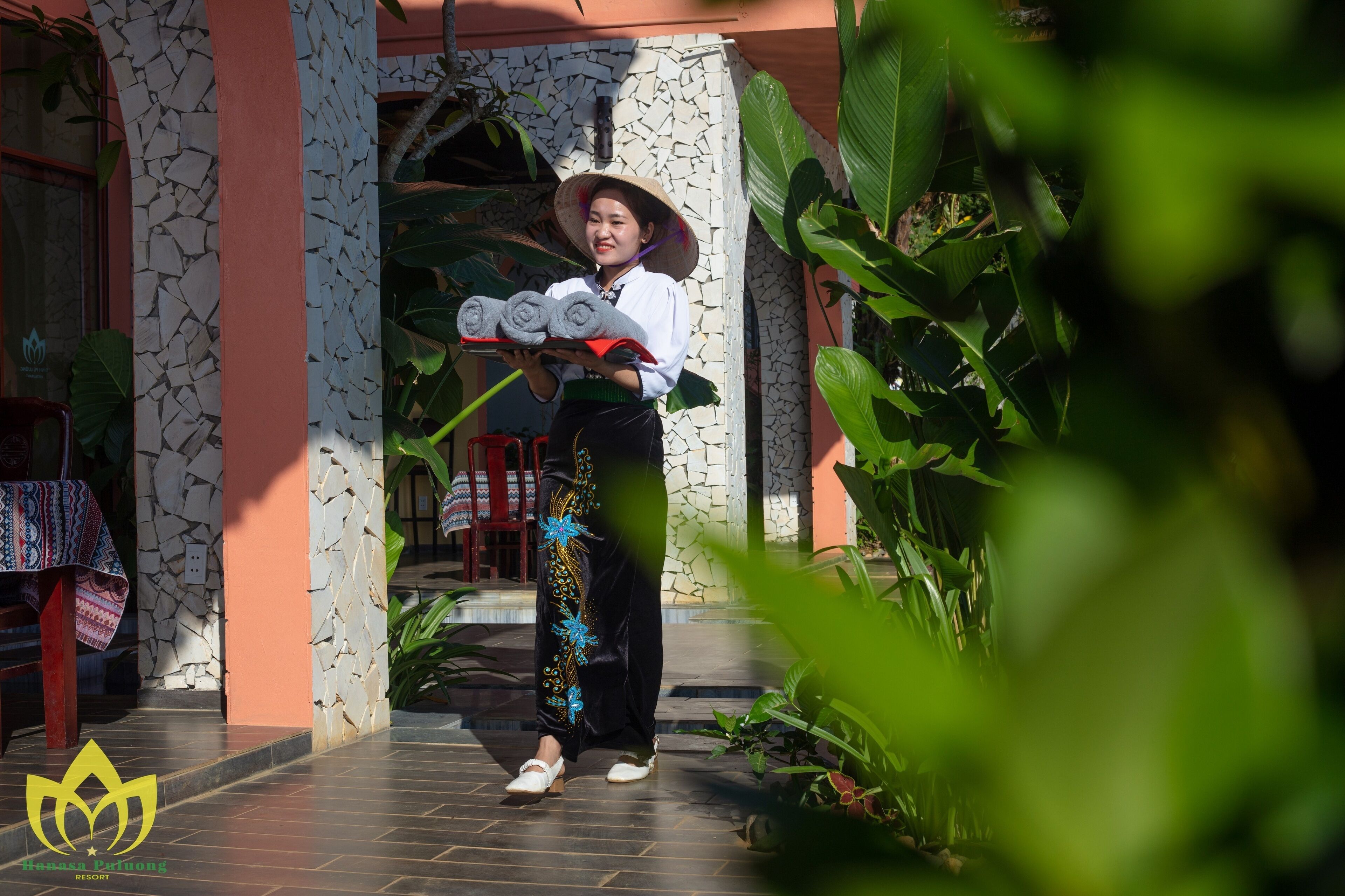 A person holding rolled towels, standing in a doorway with a stone wall and tropical plants.