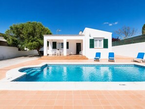 A poolside view of a white building with a patio, two blue lounge chairs, and a clear blue sky.