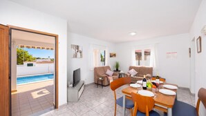 Un salon moderne avec vue sur la piscine, un coin repas avec une table dressée pour deux personnes et un téléviseur posé sur un meuble.