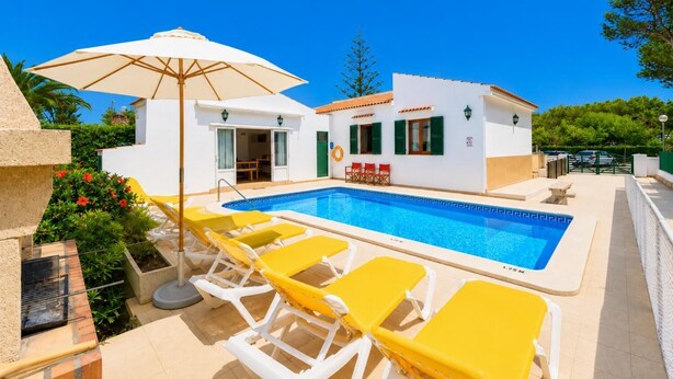 Ein Poolbereich mit Pool, gelben Liegestühlen, einem weißen Sonnenschirm und einem Gebäude mit einem Kachel­dach.