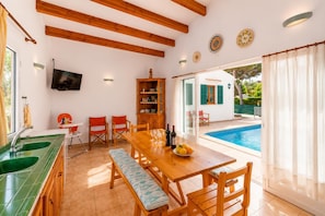 Un coin repas avec une table en bois, des chaises, un banc, une cuisine dotée d’un évier vert, et une piscine visible à travers la porte en verre.