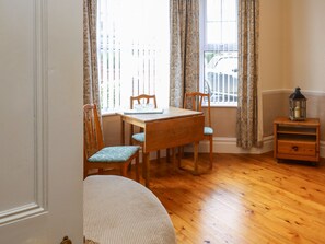 A dining area with a wooden table and chairs, a window with curtains, and a small wooden side table with a lantern. - Tidal Reach (Paignton)