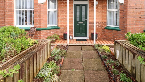 A brick house with a green door, white-framed windows, and a small porch with a checkered tile path leading to the door. - Tidal Reach (Paignton)