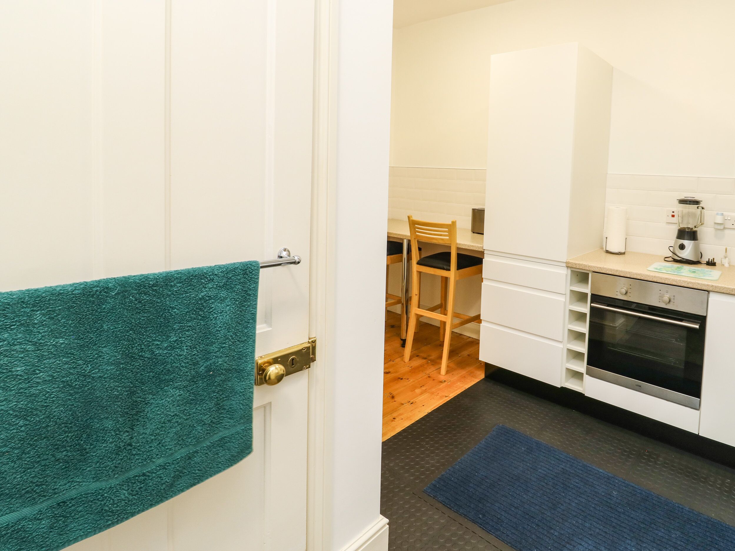 Une cuisine avec des meubles blancs, une chaise en bois, un tapis bleu et une serviette turquoise accrochée à une porte.