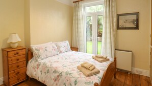 A bedroom with a wooden bed, a nightstand with a lamp, a radiator, and a window with curtains. - Tidal Reach (Paignton)