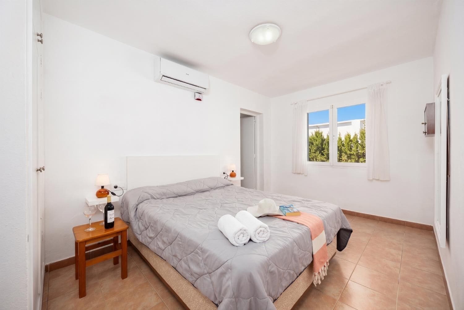 Un dormitorio con una cama, una mesita de noche con lámpara, una ventana con cortinas y un equipo de aire acondicionado.