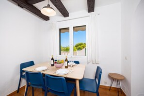 A dining area with a table set for four, blue chairs, a bowl of fruit, wine bottles, and a view of greenery through a window. - Villa Eugenia Large Private Pool, A/C, WiFi (Cala en Porter)
