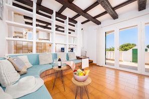 A spacious living area with a blue sofa, wooden beams, white shelving, and a balcony with a green gate. - Villa Eugenia Large Private Pool, A/C, WiFi (Cala en Porter)