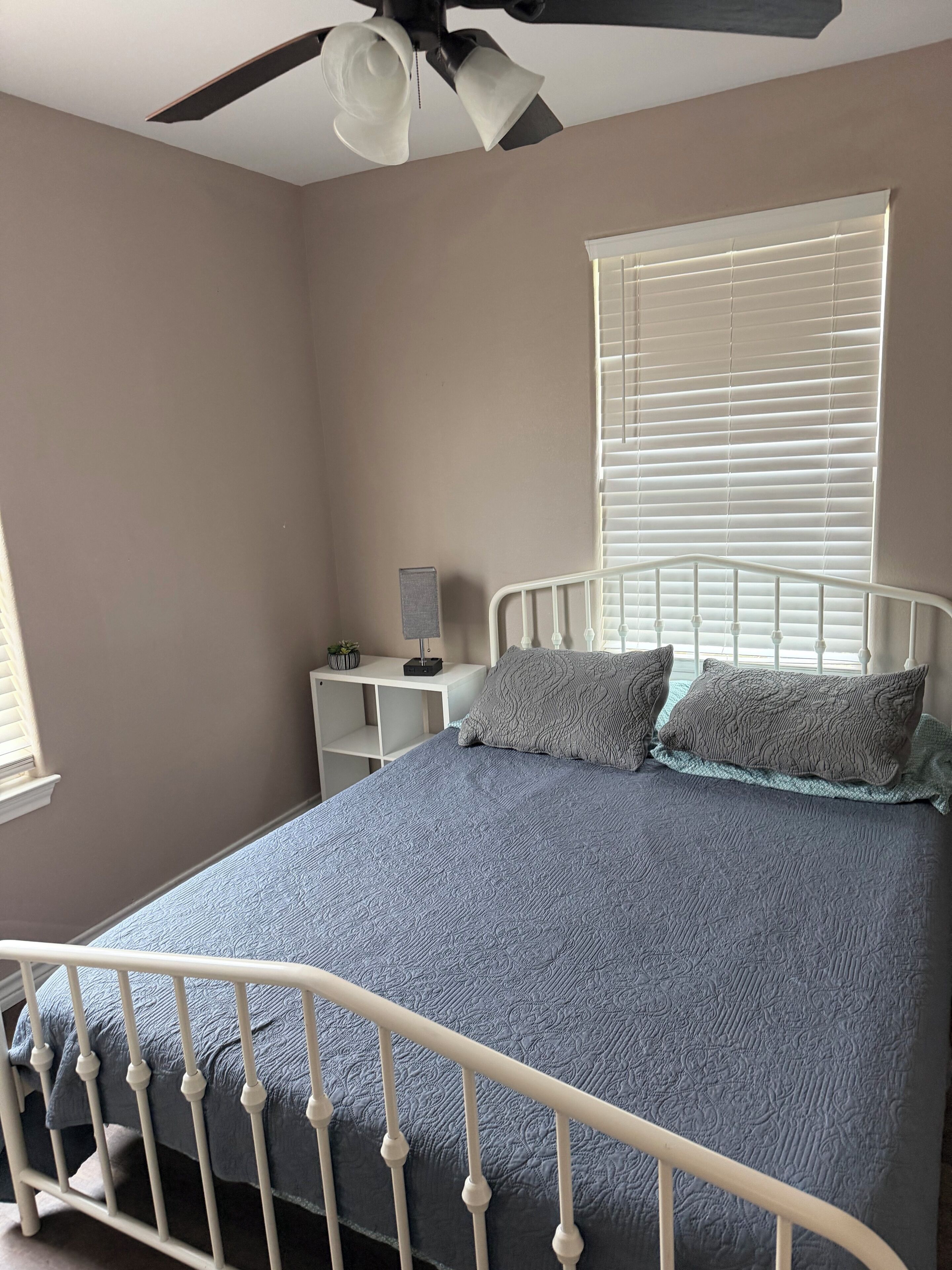 A bedroom with a bed, a nightstand, a lamp, a ceiling fan, and a window with blinds.