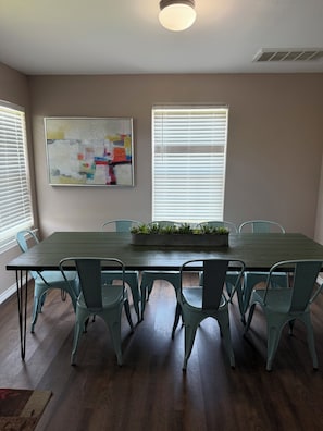 Uma sala de jantar com uma mesa comprida, seis cadeiras, uma planta em vaso e um quadro na parede.