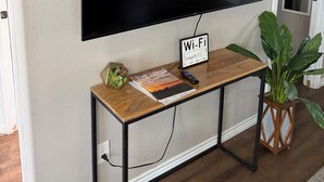 A flat-screen TV mounted on a wall, a Wi-Fi sign, a small wooden console table with a book and a remote, and a potted plant. - TRANQUILITY BAY-Spring /Summer /WINTER TEXAN FAVORITE!!  Come RELAX with us! (Aransas Pass)
