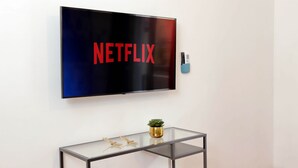 A flat-screen TV mounted on a wall with the word "NETFLIX" displayed on it. Below the TV, there is a glass table with a small potted plant and two decorative items. - Comfy apartment next to Vatican city (Roma)