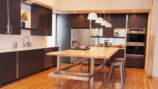 Une cuisine moderne avec des meubles de couleur sombre, un îlot central en bois, des appareils électroménagers en acier inoxydable et des luminaires suspendus.