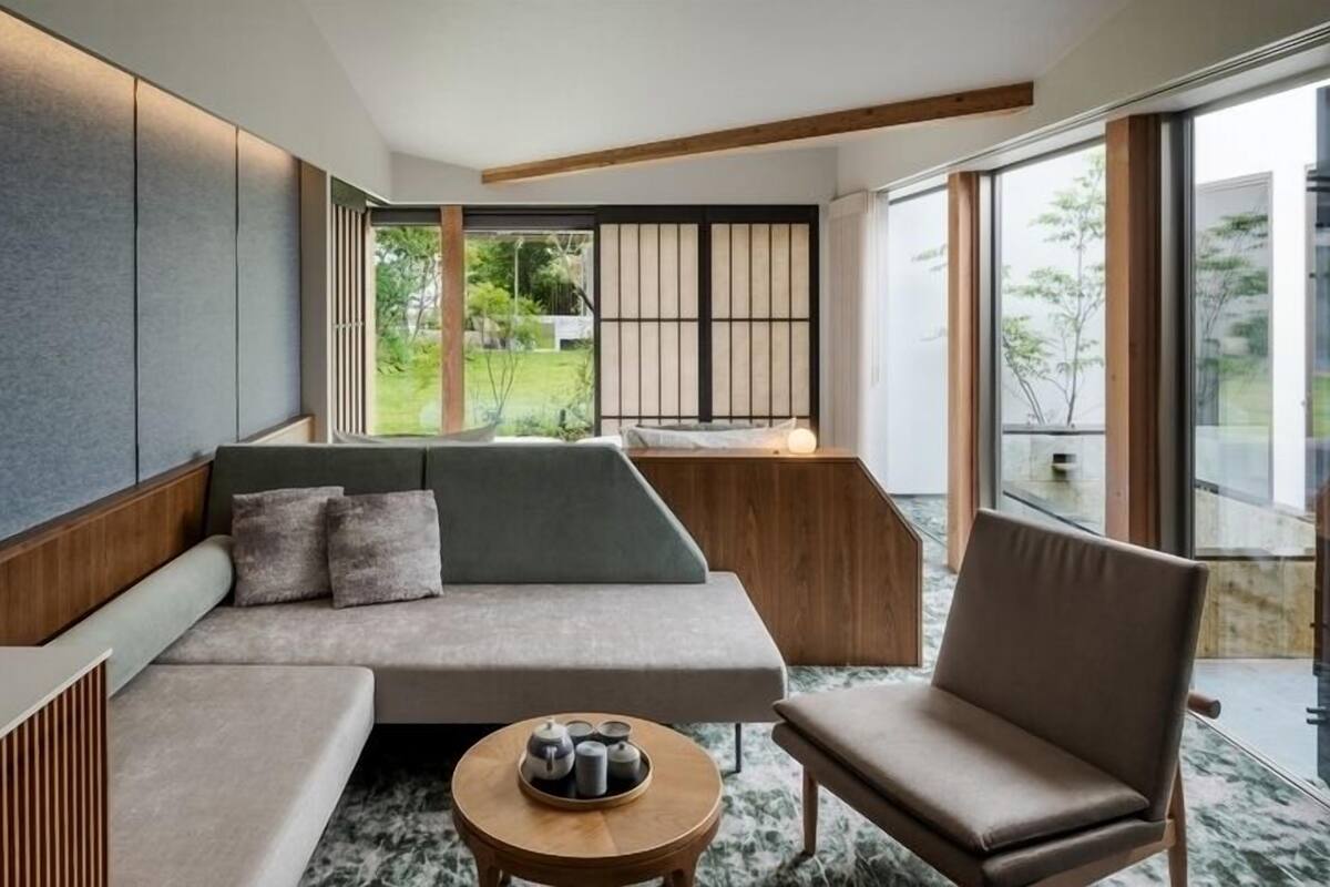 Uma sala de estar moderna com sofá, poltronas, uma mesinha com bules e grandes janelas com vista para o verde.