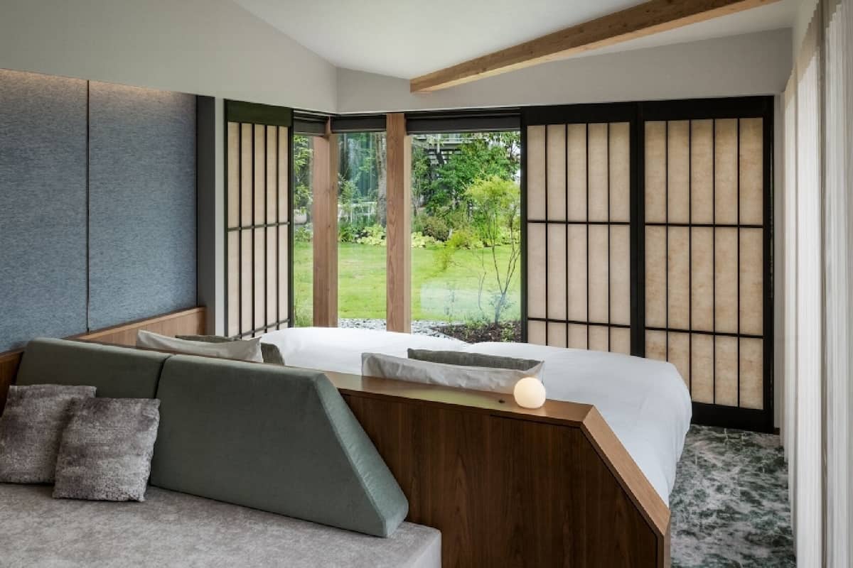 Quarto de hotel moderno com sofá, cama e vista para um jardim através de grandes janelas.