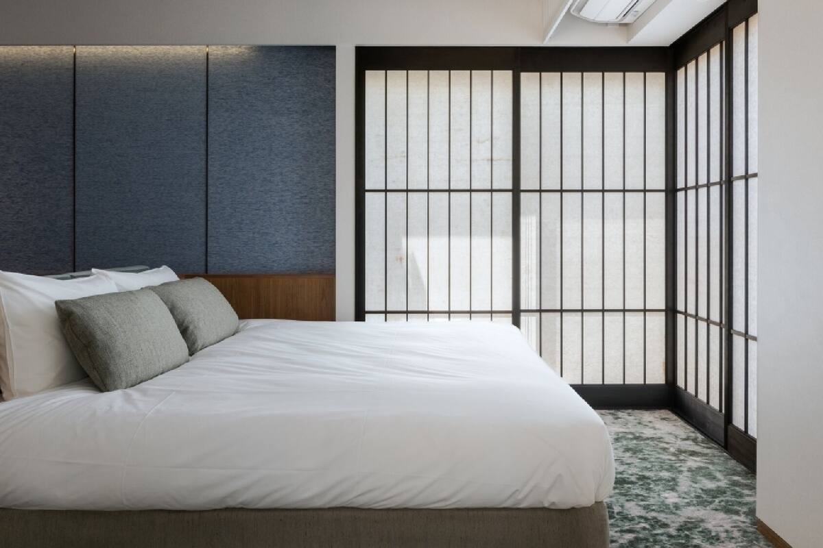 Quarto de hotel moderno com cama, mesa de cabeceira e uma janela grande com persianas verticais.