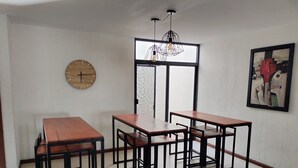 A dining area with three wooden tables and chairs, a wall clock, a framed abstract painting, and a window with a view.