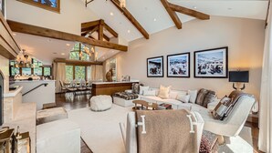 A spacious living area with a high ceiling, wooden beams, a central seating area with a sofa and ottoman, a dining area with a large table and chairs, and a kitchen with a bar.