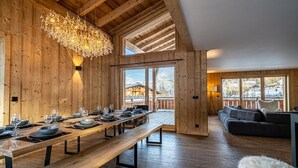 A wooden interior with a long dining table set for six, a cozy seating area, and large windows offering a view of the outside.