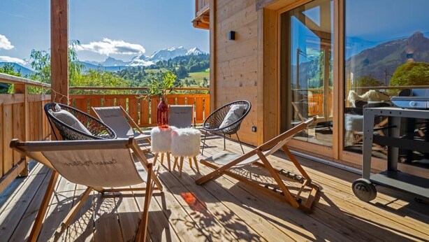 A wooden deck with two lounge chairs and a table, overlooking a mountainous landscape.