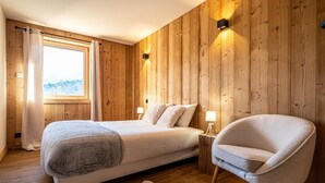A bedroom with a wooden paneled wall, a large window with curtains, a bed with pillows, a bedside table with lamps, and a chair.