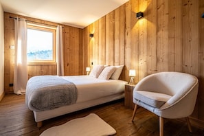 Un dormitorio con pared de paneles de madera, un ventanal con cortinas, una cama con almohadas, una mesita de noche con lámparas y una silla.