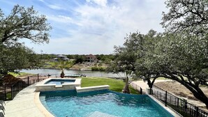 Une piscine à bord incurvé, entourée d’une clôture en métal noir, avec vue sur un lac et des maisons en arrière-plan.