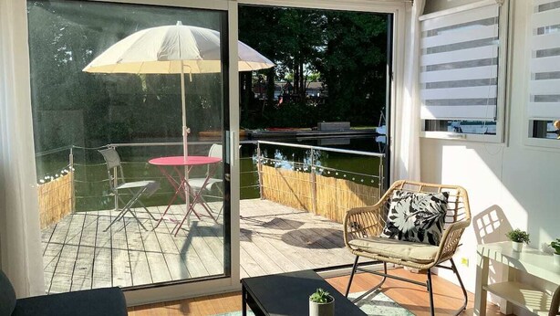 Una habitación moderna con puerta de vidrio corrediza que da a una terraza exterior. La terraza tiene suelo de madera, una mesa redonda con sillas y una sombrilla blanca. Hay una silla de mimbre con cojín y una mesita con una planta.