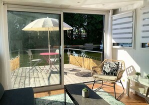A modern room with a sliding glass door leading to an outdoor deck. The deck has a wooden floor, a round table with chairs, and a white umbrella. There is a wicker chair with a cushion and a small table with a plant.