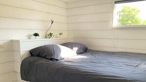 Un dormitorio con cama, paredes blancas, una ventana y una planta pequeña en la mesita de noche.