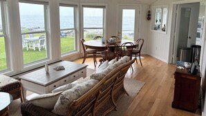 A room with a large dining table and chairs, a bench with cushions, and a view of the ocean through large windows.