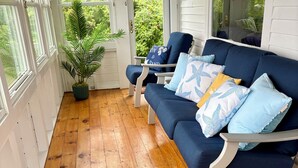 A covered porch with a wooden floor, a blue sofa with patterned cushions, and a potted plant.