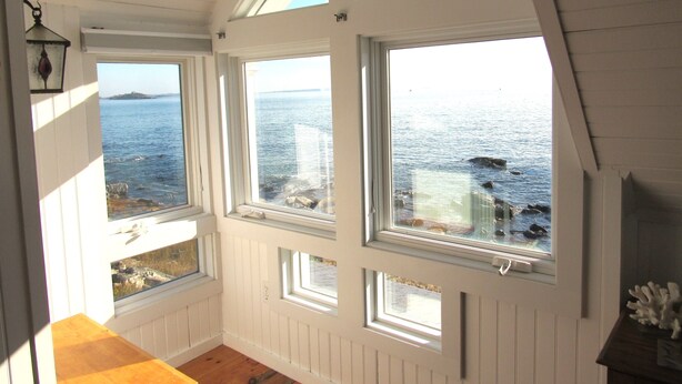 A room with a view of the sea through multiple windows, wooden paneling, and a wooden floor.