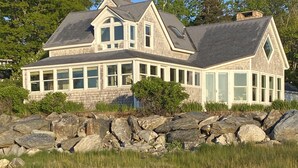 A large, two-story house with a stone facade, multiple windows, and a prominent chimney. It is situated on a rocky shoreline with a view of the surrounding landscape.