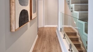Un pasillo con piso de madera, una escalera con barandilla de cristal y cuadros en las paredes.