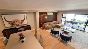 Una sala moderna con un sofá seccional, una mesa de comedor con sillas, un televisor montado en la pared y un ventanal que ofrece vistas al exterior.