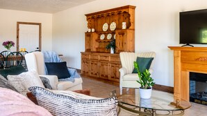 A living room with a sofa, armchairs, a glass coffee table, a wooden cabinet with decorative plates, and a fireplace.