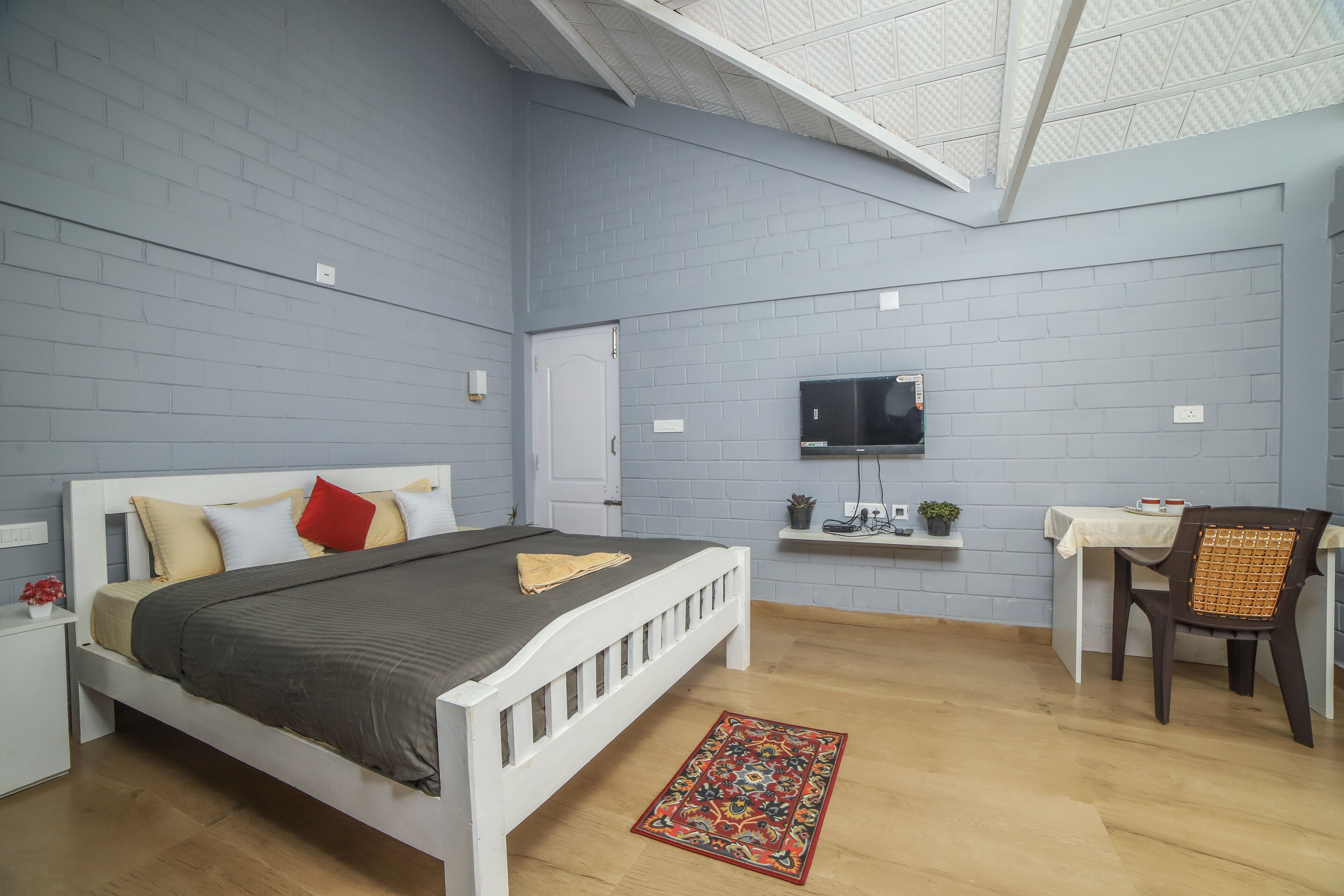 A bedroom with a bed, a desk, a chair, a TV, and a small shelf with a plant and shoes.