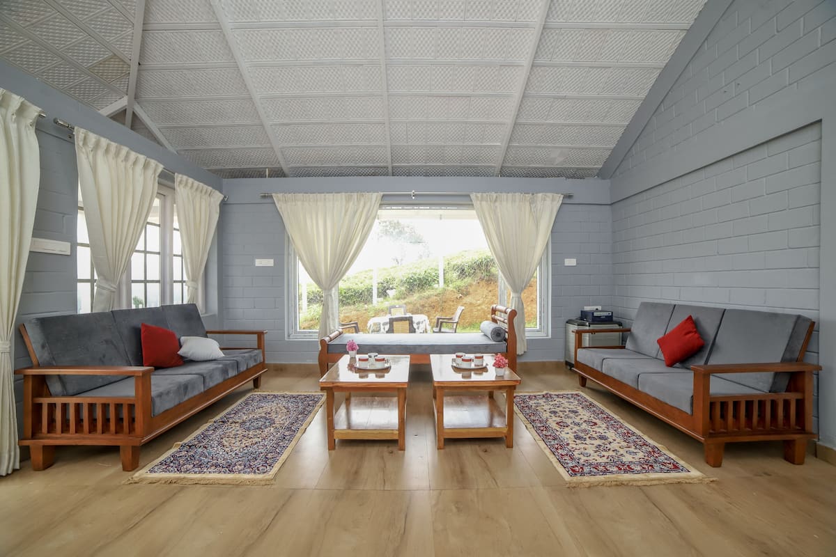 A spacious living room with two sofas, a coffee table, and a bench. Large windows offer a view of greenery outside.