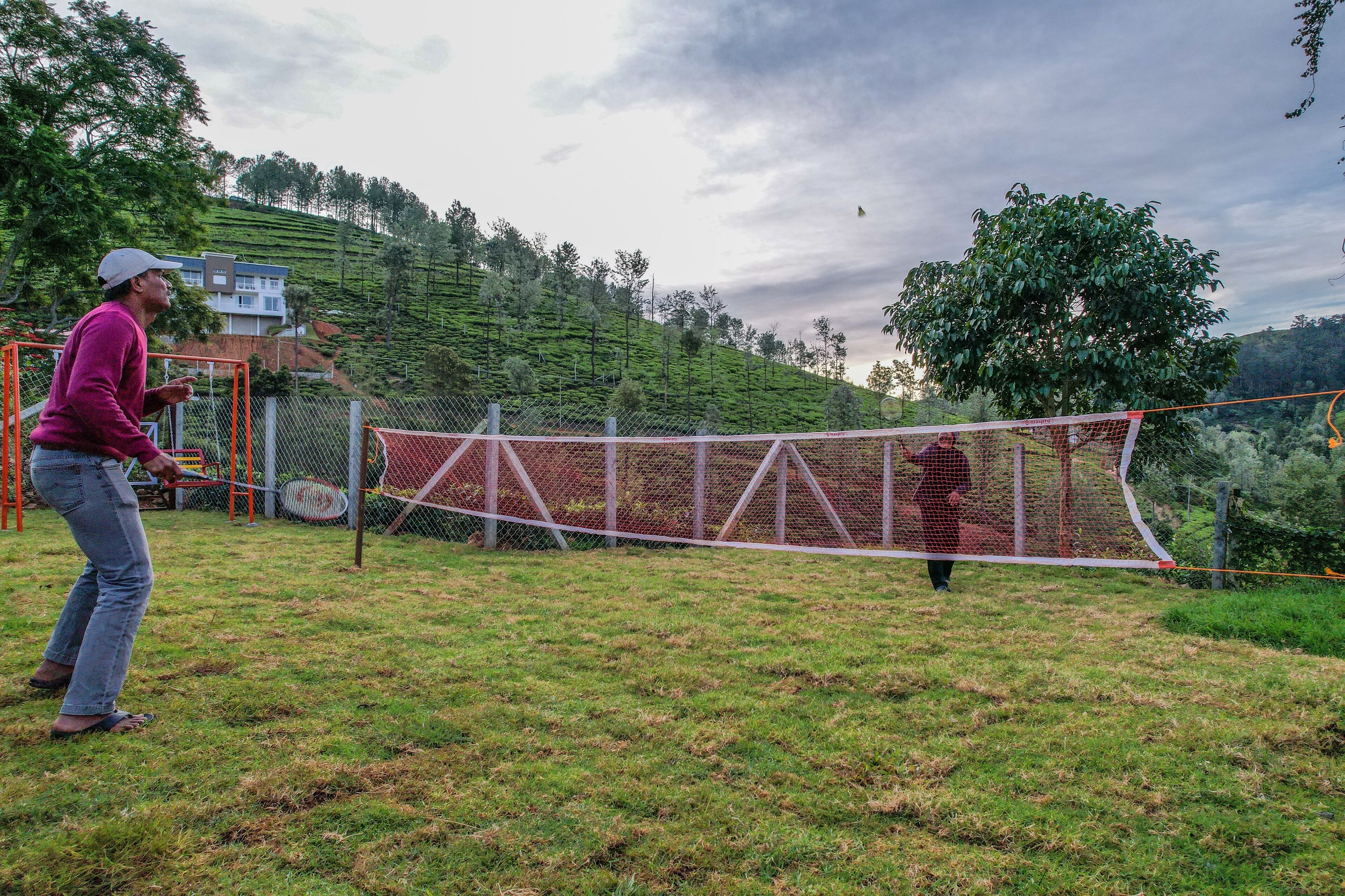 A person playing badminton in an outdoor setting with a net, surrounded by greenery and a hilly landscape.