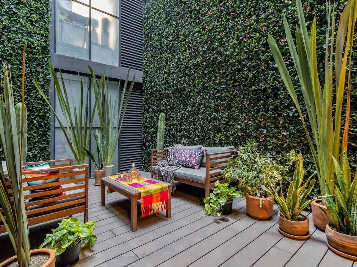 Una terraza de madera con un sofá y una mesa, rodeada de plantas en macetas y un muro verde.
