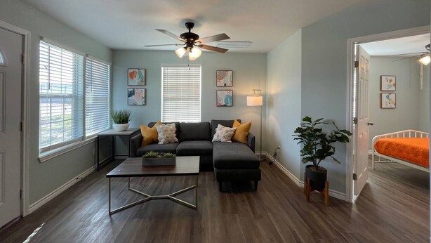 A living room with a dark grey sofa, a coffee table, and a ceiling fan. There are two windows with blinds, a plant, and artwork on the walls.