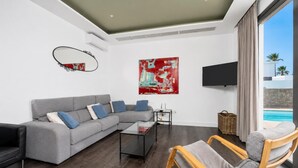 A modern living room with a grey sectional sofa, a glass coffee table, a wooden rocking chair, a flat-screen TV, and a large abstract painting.