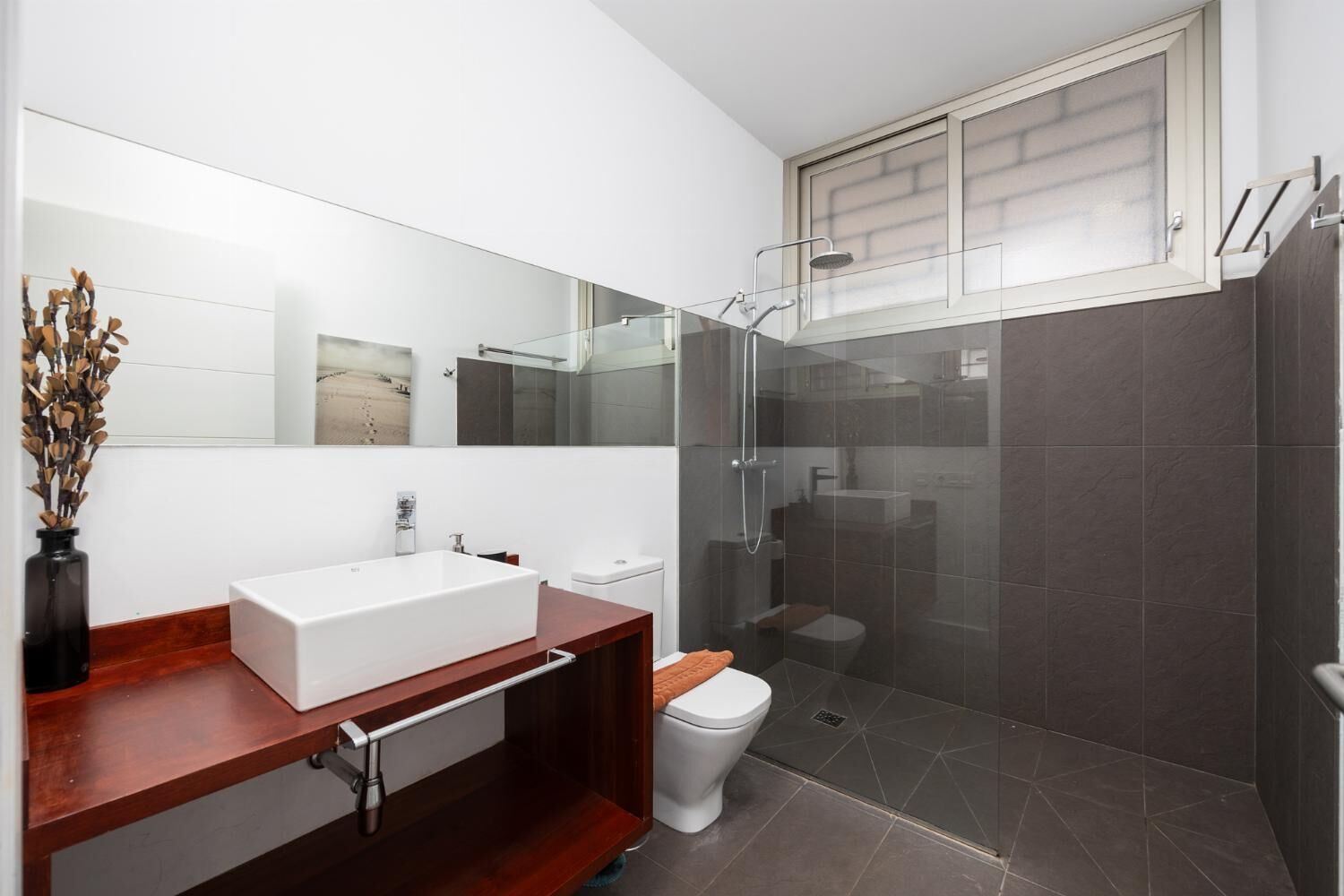 Ein modernes Badezimmer mit einem großen Spiegel, einem weißen Waschbecken, einer Holzausstattung, einer Toilette und einer Duschkabine aus Glas.