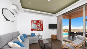 A modern living room with a grey sofa, a glass coffee table, a wooden rocking chair, a TV mounted on the wall, and a large abstract painting.