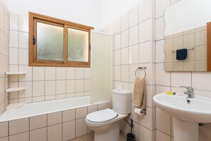 Une salle de bain avec des toilettes, un lavabo et une baignoire. Il y a une fenêtre au-dessus des toilettes et un porte-serviettes sur lequel est accrochée une serviette.
