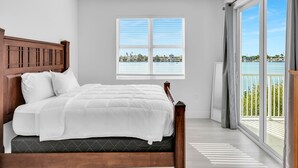 Quarto com cabeceira de madeira, cama com roupa de cama branca, ventilador de teto e janela com vista para um corpo de água e edifícios.