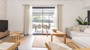 A modern living room with a sofa, a TV, a dining area with chairs and a table, and a balcony with outdoor furniture.